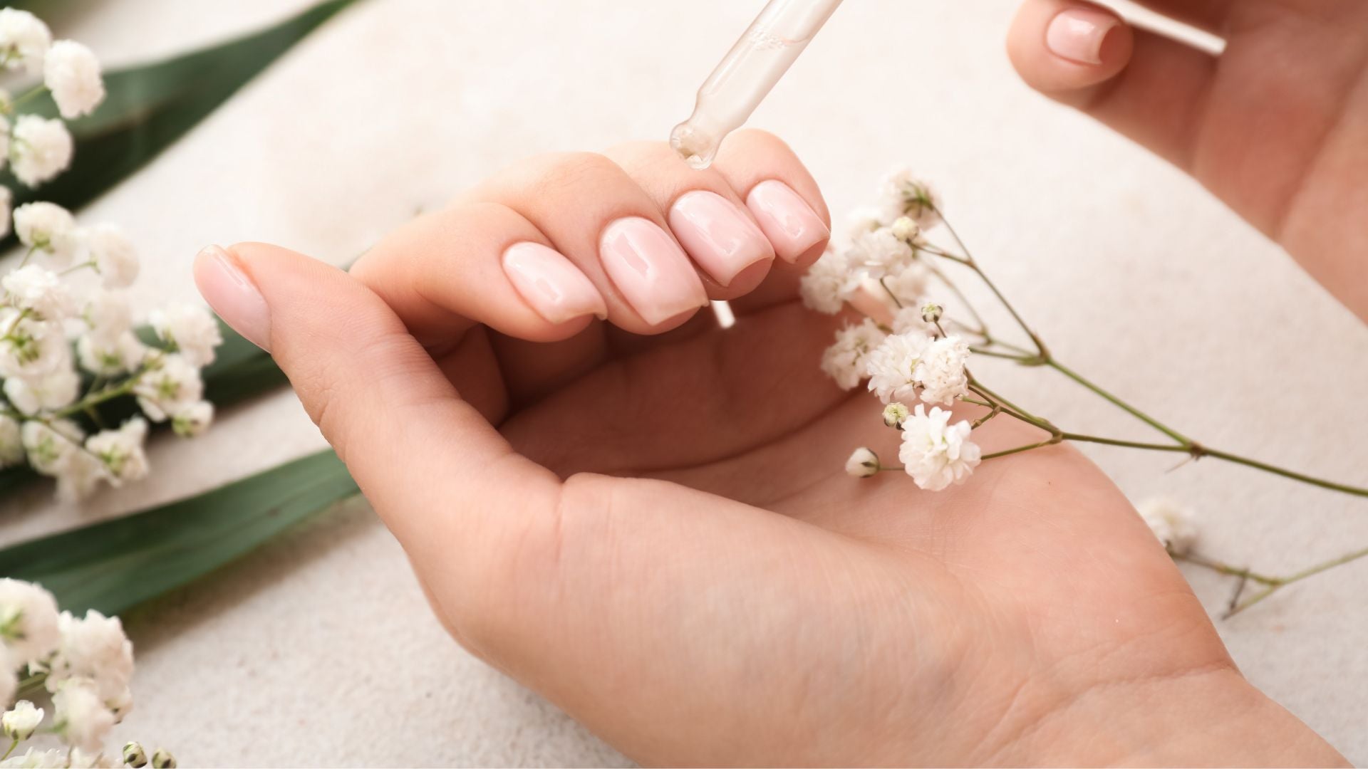 Gocce di olio naturale applicate su mani e unghie curate, fiori bianchi, trattamento delicato e rigenerante