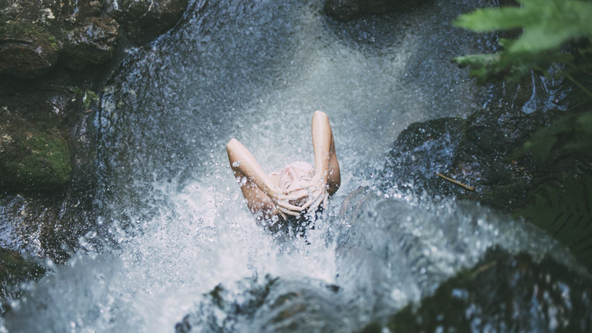 Donna sotto una cascata naturale immersa nella freschezza dell'acqua, simbolo di pulizia profonda e rigenerazione ispirata ai detergenti corpo di Rugiada Mediterranea